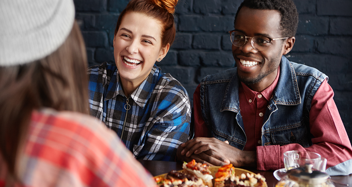 Indoor shot of interracial group of three young stylish people having easy and lively conversation while relaxing at modern cafe, eating their lunch, talking and laughing at each other's jokes.